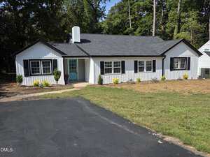 Charming single-story house at 115 Red Bud Drive, Henderson, with white siding, black trim, and a gray shingled roof. Features a turquoise front door, small shrubs along the facade, a paved driveway, and trees in the background.