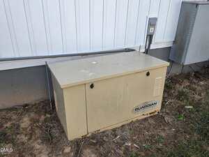 A beige Guardian generator sits on the ground next to the exterior wall of a building, near an electrical box and a meter on a patch of dirt and grass.