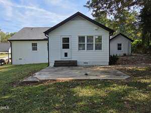 A small, white, single-story house with black trim is shown from the back. There is a concrete pad, a few steps leading to a door, multiple windows, and a grassy yard with scattered leaves and trees in the background.