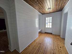 A bright hallway with white shiplap walls, a wood plank ceiling, and light wood floors. There is a door with glass panes at the end, letting in natural light, and a small bench area to the left of the door.
