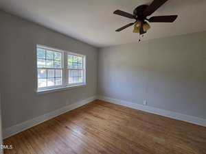 An empty room with light gray walls, a wooden floor, a ceiling fan with light fixtures, and a large window letting in natural light. The space is unfurnished and appears clean and well-maintained.