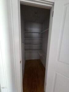 A small, empty walk-in closet with white walls, white shelves on the left and back walls, a hanging rod on the right, and a wooden floor. The door is partially open.