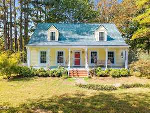 Located at 8707 Grassy Creek Road in Bullock, this yellow house with a green roof features white trim, a red front door, and a covered front porch with columns and steps leading to a stone walkway amid grass, bushes, and tall trees.