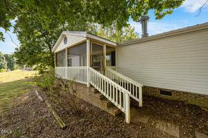 A single-story house with white siding features a screened-in porch and a ramp with railings leading to the entrance. The yard has some plants and mature trees, and the ground is covered in leaves and mulch.