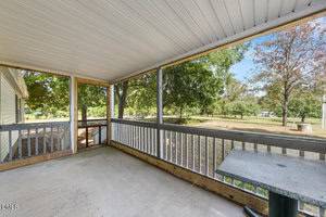 Screened-in porch with a concrete floor, white ceiling, and wooden railings, overlooking a grassy yard with trees and a blue sky visible in the background. A stone table stands on the right side.