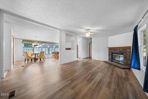 Open living room and dining area with wood-style flooring, white walls, a stone fireplace, ceiling fan, large windows with blue curtains, and a dining table set near sliding glass doors.