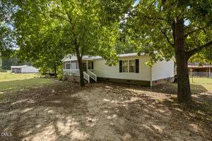 A single-story white manufactured home with a brick foundation sits beneath large leafy trees. There is a small front porch with railings, and the yard has sparse grass and patches of bare earth.