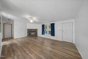 A spacious living room with wood-look flooring, a stone fireplace with a wood mantel, a ceiling fan, white walls, and a window with blue curtains next to a white front door.