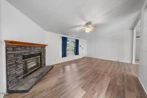 Living room with light wood flooring, white walls, a stone fireplace with a wood mantel on the left, a ceiling fan with light in the center, and a window with blue curtains next to a white front door.