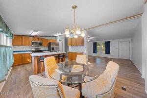 A kitchen and dining area with wood cabinets, stainless steel appliances, a central island, and a glass-top table with four patterned chairs. The room features wood flooring and light fixtures above the table.