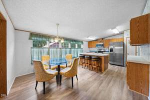A kitchen and dining area with wood flooring, a round glass dining table with four yellow upholstered chairs, a kitchen island with three stools, wood cabinets, stainless steel appliances, and large windows with green curtains.