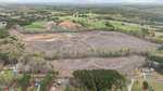 Aerial view of a large, cleared plot of land surrounded by trees and rural homes, with patches of green fields and forest in the background under a cloudy sky.