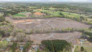 Aerial view of a large, cleared plot of land surrounded by trees and rural homes, with patches of green fields and forest in the background under a cloudy sky.