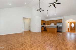 Spacious open-concept room with wood floors, high vaulted ceiling, two ceiling fans, a kitchen with wooden cabinets, stainless steel appliances, and an island; entryway with double doors visible in the background.