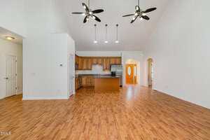 Spacious open-concept kitchen and living area with wood flooring, wood cabinets, an island, high ceilings, three black ceiling fans, and pendant lights. White walls and an arched entryway are visible.