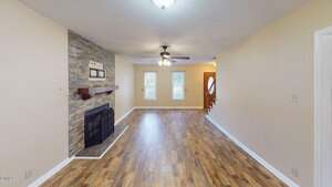 A living room with wood flooring, beige walls, a ceiling fan, and overhead light. There is a stone fireplace with a wooden mantel on the left and two windows near a wooden front door with stairs beside it.