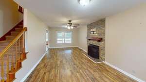 A living room with wood-look flooring, a staircase on the left, a ceiling fan with lights, a fireplace with a stone surround and wooden mantel, and a window at the far end letting in natural light.