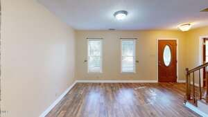 Empty room with light-colored walls, two windows with blinds, wooden front door with an oval glass insert, wood-look flooring, and a wooden staircase railing on the right. Ceiling light fixture is turned on.
