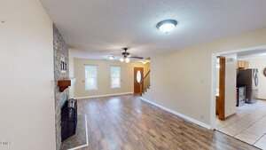 A living room with wood-look flooring, a stone fireplace on the left, light-colored walls, ceiling fans with lights, and windows at the far end. An open doorway leads to a kitchen with tile flooring and visible appliances.