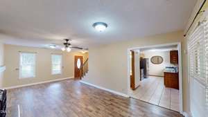 A spacious living area with wood flooring, beige walls, and two ceiling lights; an open doorway leads to a kitchen with tile flooring and wooden cabinets. Three windows and a staircase are visible.