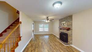 A living room with wood flooring, beige walls, a ceiling fan, a staircase with wooden railing on the left, and a fireplace with a stone surround and wooden mantel on the right wall. Large window at the back.