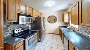 A galley-style kitchen with wood cabinets, stainless steel appliances, tile backsplash, double sink, and tiled floor. A round mirror hangs on a beige wall at the far end, reflecting a tripod and camera.