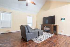 A living room with hardwood floors, a gray armchair with an ottoman on a patterned rug, a large flat-screen TV on a wooden stand with a fireplace, and two windows with blinds on light-colored walls.