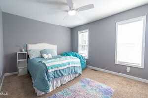 A neatly made bed with blue and turquoise bedding sits against a gray wall in a bedroom. There is a white nightstand, a bean bag chair, two windows, beige carpet, and a ceiling fan with lights.