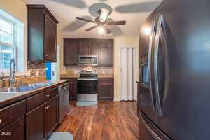 A kitchen with dark wood cabinets, stainless steel appliances, a double sink under a window, a black refrigerator, electric stove, microwave, ceiling fan, laminate wood flooring, and beige walls.