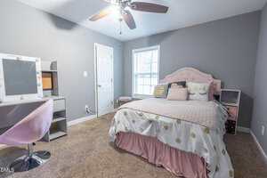 A bedroom with gray walls, beige carpet, a bed with pink and white bedding, a makeup vanity with a mirror and chair, a window with blinds, and a ceiling fan with lights.