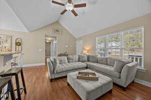 A living room with a gray L-shaped sectional sofa, matching ottoman, wooden floors, a ceiling fan, neutral walls, and large windows letting in natural light. A hallway and part of a kitchen are visible in the background.