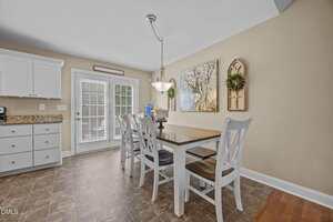 A kitchen dining area features a wooden table with four white chairs, a hanging light above, beige walls with framed art and a window decor piece, and a door with blinds leading outside. White cabinets and granite countertops are visible.