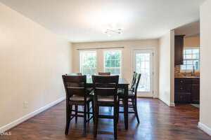 A dining area with a dark wooden table and six matching chairs on a hardwood floor, two large windows, and a glass door leading outside. The adjacent kitchen is partially visible on the right.