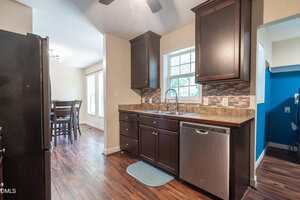 A kitchen with dark wood cabinets, a stainless steel dishwasher and sink, brown countertops, a small blue mat, and wood-style flooring. A dining area with a table and chairs is visible in the background.