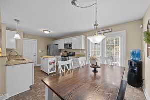 A kitchen with white cabinets, stainless steel appliances, and a wooden dining table with four chairs. There is a water dispenser in the corner and pendant lighting above the table and sink. Sunlight streams in through window blinds.