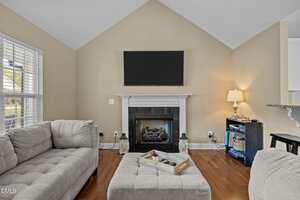 A living room with beige walls, a mounted flat-screen TV above a white fireplace, gray sectional sofas, a cushioned ottoman with a tray, wood flooring, a window with blinds, and a small lamp on a cabinet.