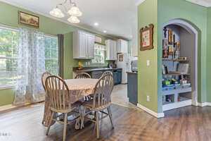A dining area with a round table covered by a floral tablecloth and four wooden chairs. The room has green walls, wood flooring, large windows with sheer curtains, and an adjacent kitchen and arched doorway with shelves.