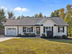 Charming single-story house in Stem at 415 Angus Place with white siding, black shutters, an attached garage on the left, a covered front porch with chairs, trimmed bushes, and a well-maintained front lawn under a partly cloudy sky.