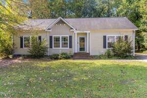 Single-story beige house with white trim and dark shutters, featuring a central front door with small stoop and steps, surrounded by green grass, bushes, and trees in the background.