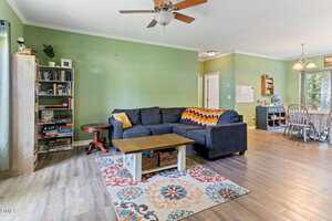 A living room with green walls, a dark sectional sofa, a colorful zigzag throw, a wooden coffee table on a patterned rug, a bookshelf with books and board games, and a dining area visible in the background.