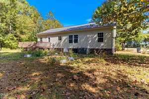 A single-story beige house with white trim sits in a yard with scattered leaves. The backyard has a wooden deck and several metal garden beds. Trees and grass surround the property under a clear blue sky.