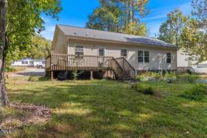 A single-story beige house with a dark roof and a wooden deck sits in a grassy yard with trees, shrubs, and a neighboring house visible in the background under a clear blue sky.