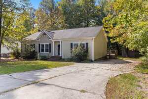 A single-story beige house with white trim, a gabled roof, and a brick front porch sits beside a concrete driveway. The yard is partially shaded by tall trees with green leaves. Shrubs line the front of the house.