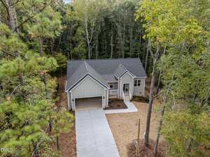 A single-story house with gray siding and a front porch sits among tall trees. The home has a gabled roof and a driveway that leads to a covered carport. The yard is partially landscaped with mulch and grass.