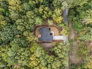 Aerial view of a single house with a gray roof, surrounded by dense green trees. The house has a driveway leading to a road, and the yard is mostly cleared with some brown patches.
