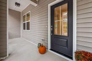 A tan house with white trim features a black front door with glass panels. A pumpkin and a decorative gourd sit on the porch near the door. A potted plant is at the edge of the image.