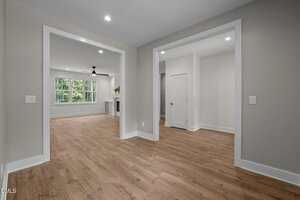A modern interior space with light wood flooring, light gray walls, white trim, recessed ceiling lights, and large windows allowing natural light into the living area. A ceiling fan and fireplace are visible in the background.