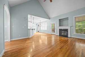 A spacious living area with light blue walls, vaulted ceiling, hardwood floors, a white fireplace, and large windows. An open kitchen with pendant lights and an arched entryway is visible to the left.