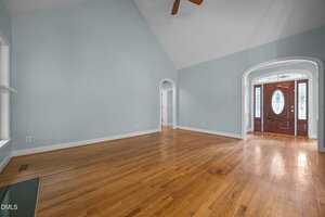 A spacious, empty living room with light blue walls, hardwood floors, a vaulted ceiling with a ceiling fan, and an arched entryway leading to a double front door with decorative glass panels.