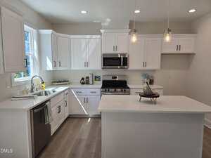 Modern kitchen with white cabinets, stainless steel appliances, a center island, pendant lights, and light wood flooring. A window above the sink lets in natural light and decorative items are placed on the counters.